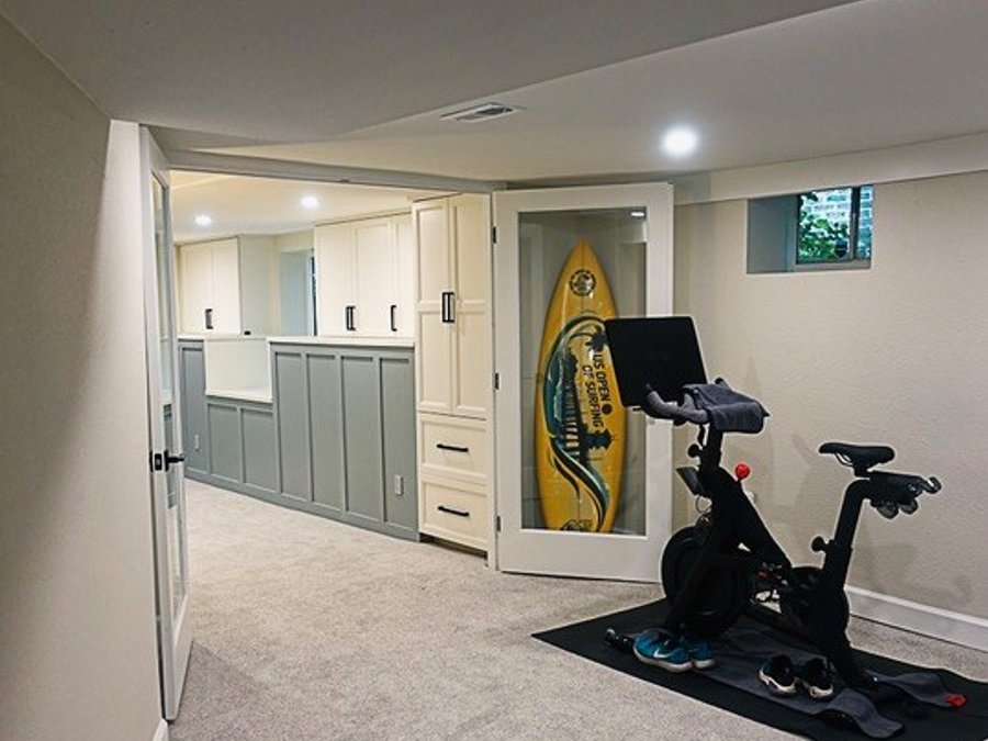 Finished basement home gym with Peloton, surfboard storage cabinet, and gray wainscoting