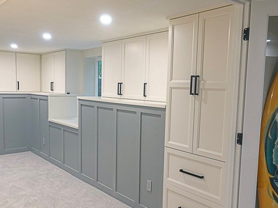 Basement built-in storage zone with white shaker cabinets, gray board-and-batten wainscoting, and farmhouse sink