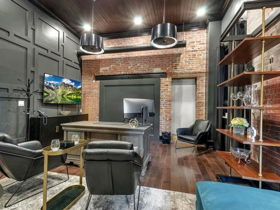 Home office with dark paneling, exposed brick, gold shelving, and leather chairs